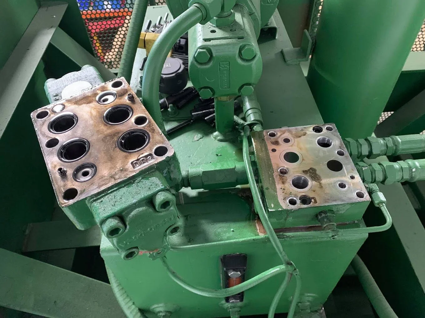 A green hydraulic machine with two metal plates, each with multiple circular openings, some filled with black rubber seals, connected by green piping.