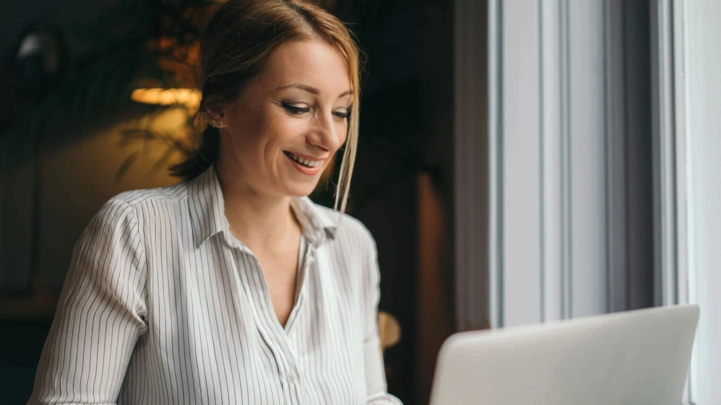 A woman with blonde hair smiling while looking at a laptop in a cozy, well-lit room near a window.