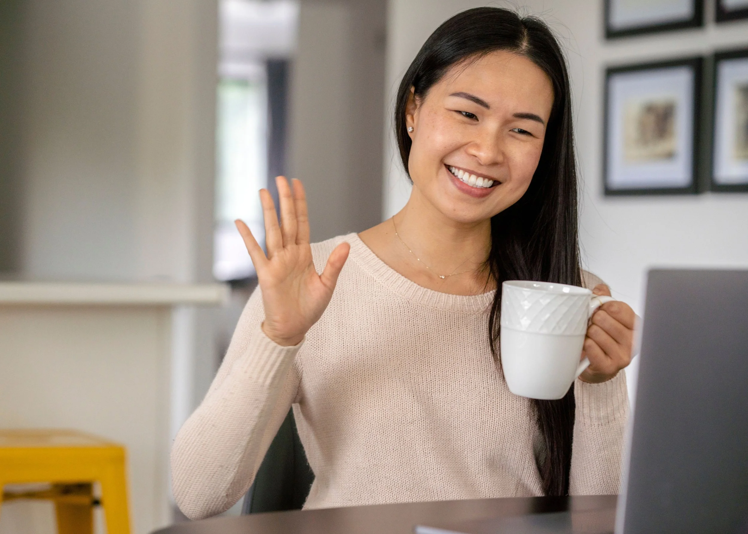 A woman with long black hair smiling and waving while holding a white mug, sitting at a desk with a laptop in front of her.