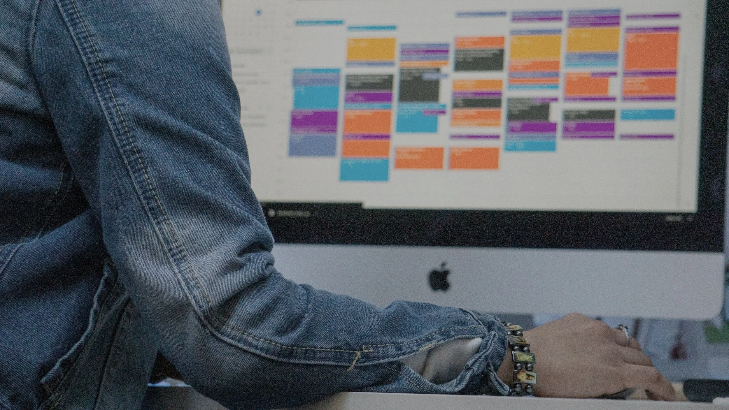 A person wearing a denim jacket working on an iMac computer with a colorful, blurred calendar or schedule on the screen.