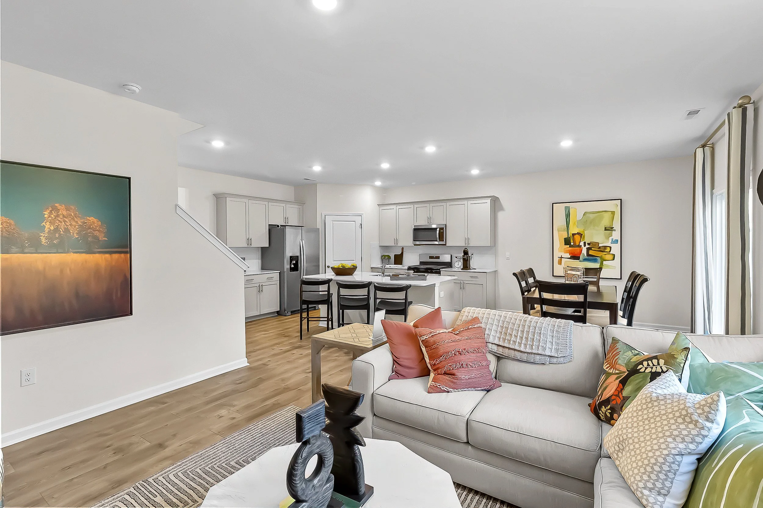 Open-concept living room and kitchen with white walls, hardwood flooring, and modern furniture, including a white sofa with decorative pillows, a wooden dining table, and kitchen with light gray cabinets and stainless steel appliances.