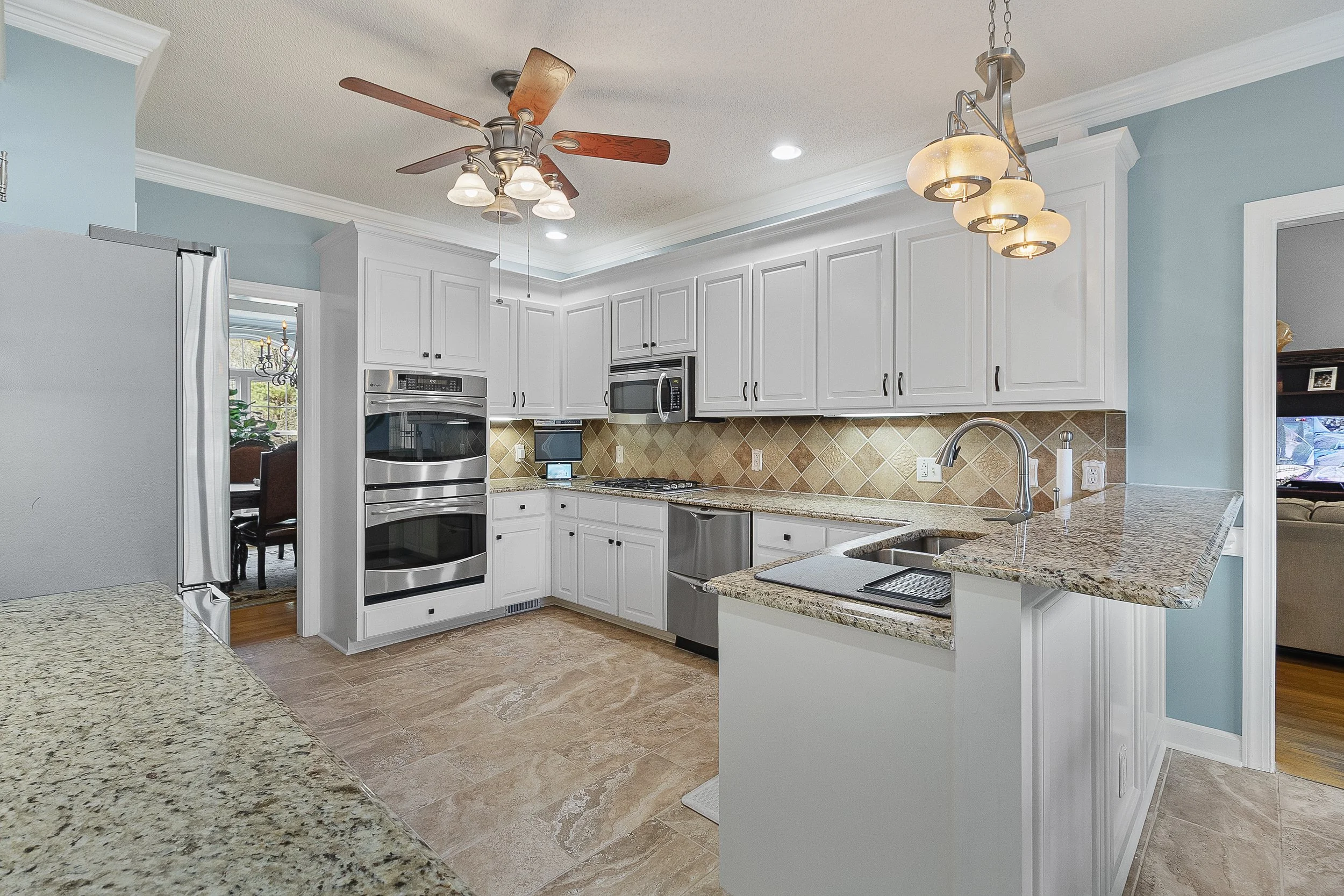 Modern kitchen with white cabinets, granite countertops, stainless steel appliances, tiled backsplash, and a breakfast bar. Ceiling with light fixtures and fan, adjacent to dining and living areas.