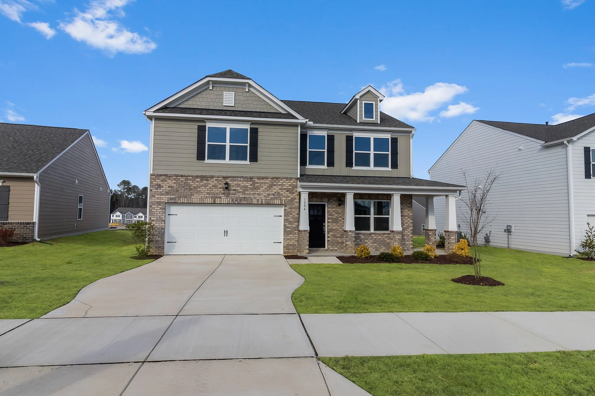 Front view of a modern two-story house with a brick and siding exterior, white garage door, front porch with white columns, and a well-maintained lawn with small shrubs and a young tree under a blue sky with some clouds.