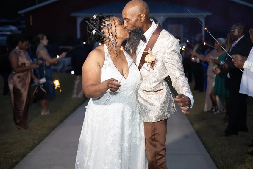 A couple has a romantic moment kissing at their wedding reception, holding sparklers, with friends and family in the background.