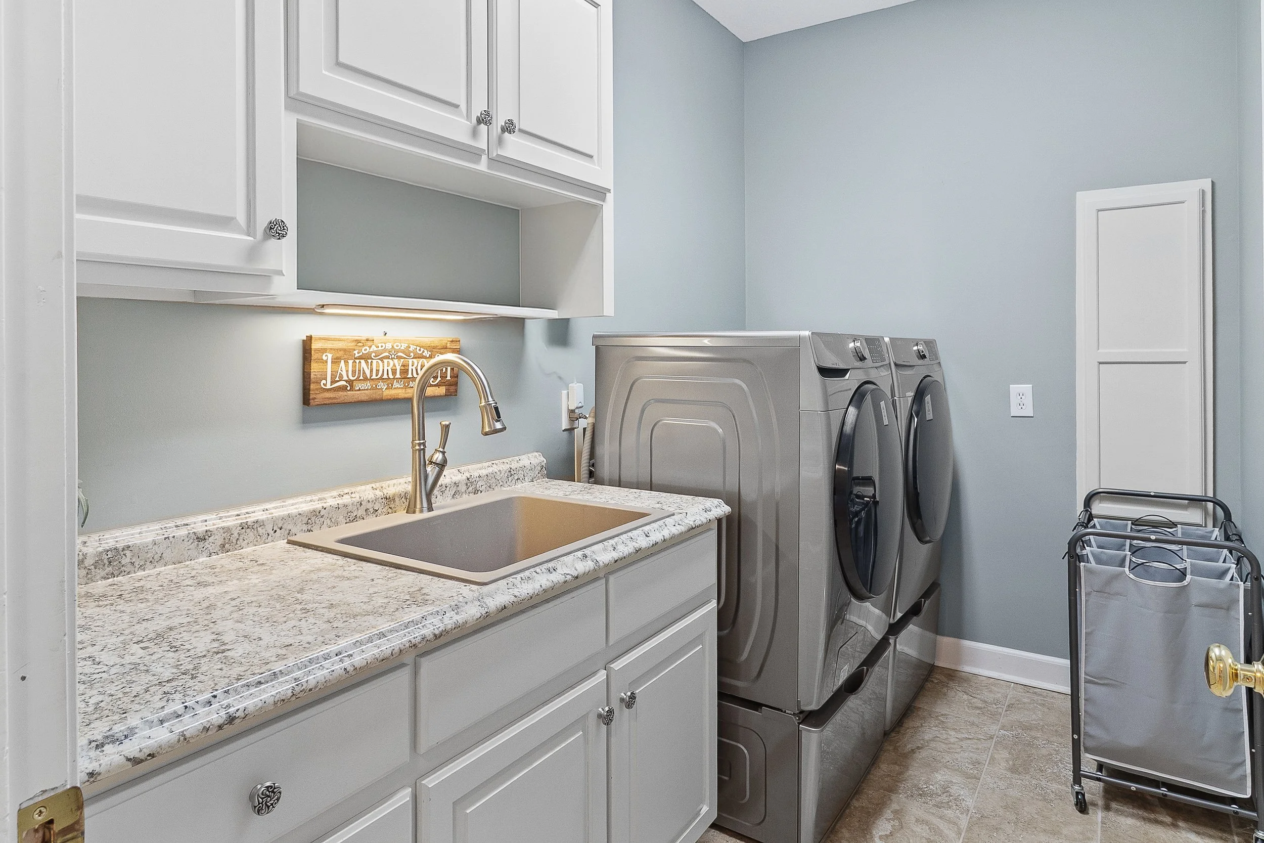 A laundry room with a white cabinet, a granite countertop with a sink, a gold faucet, a wooden sign that says 'Loads of Fun Laundry Room,' and a gray washing machine and dryer.