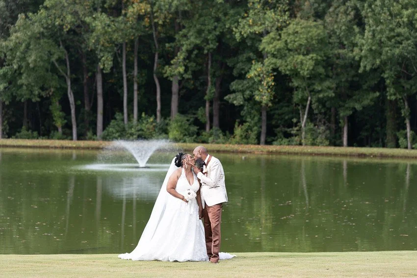 A bride and groom kissing near a pond in a park, with a fountain in the background and trees surrounding the water.