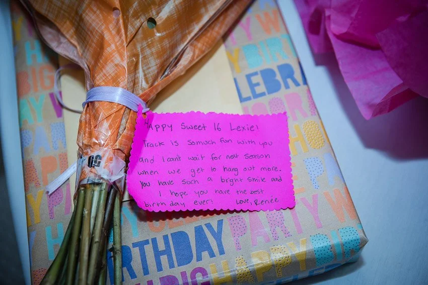 A wrapped birthday gift box decorated with colorful birthday-themed paper, a pink note card with a handwritten birthday message, and a yellow and orange bouquet of flowers.
