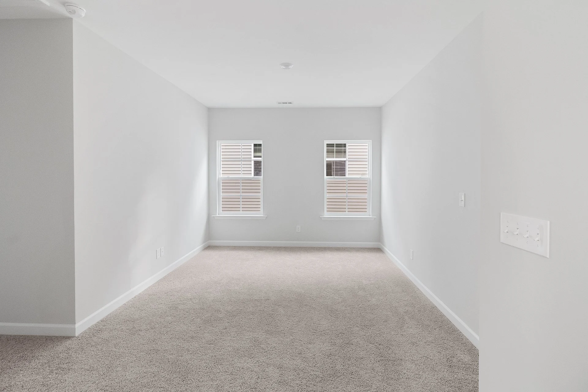 Empty room with white walls, beige carpet, and two windows with blinds, no furniture.