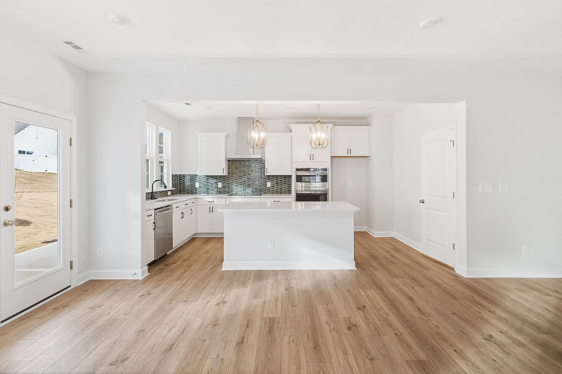 Empty modern kitchen with white cabinets, stainless steel appliances, and wooden flooring, with a door to the outside on the left.