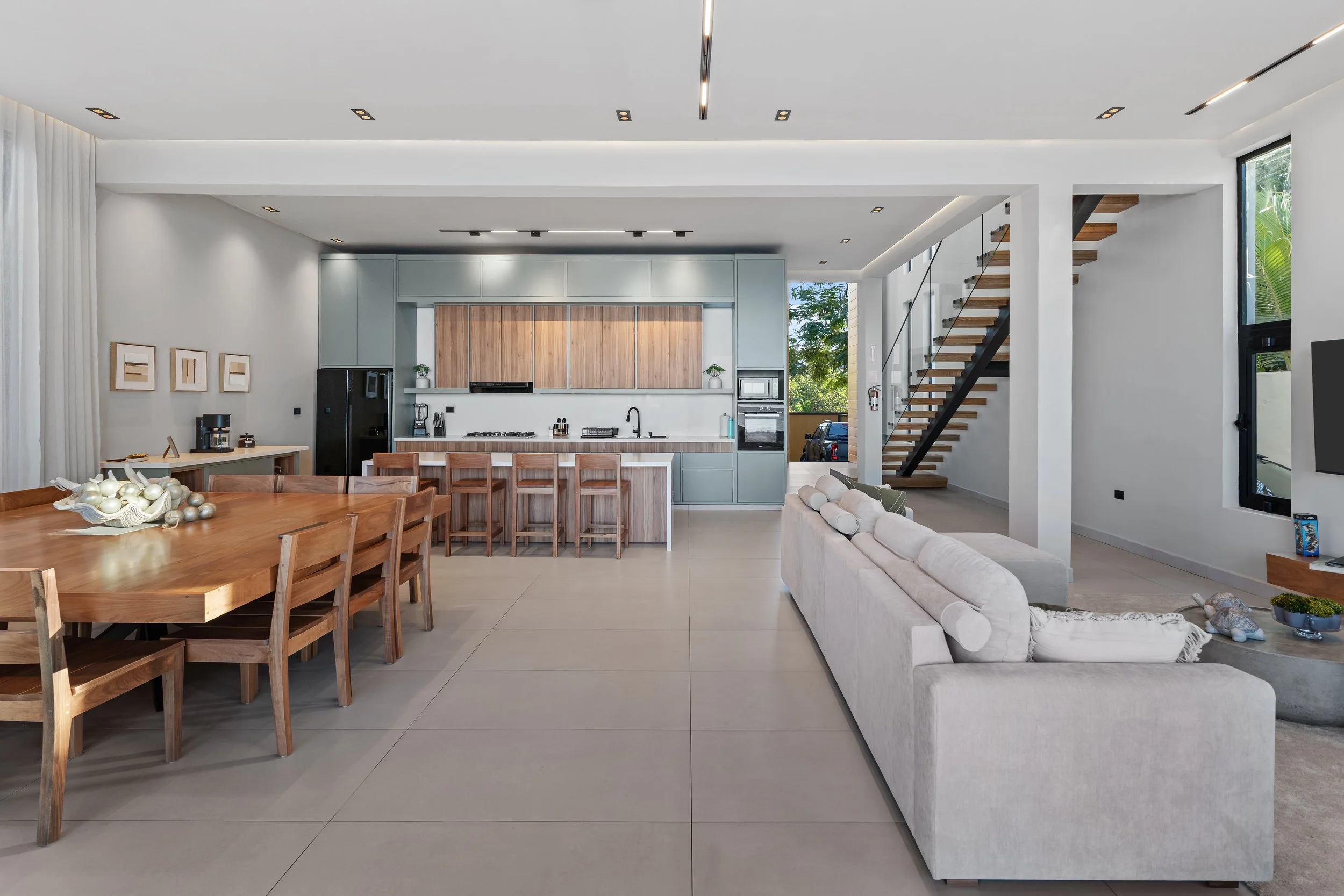 Open-concept living room and kitchen with modern decor, dining table with chairs, white sofa, staircase with wooden steps and black railing, large windows, and minimalistic style.