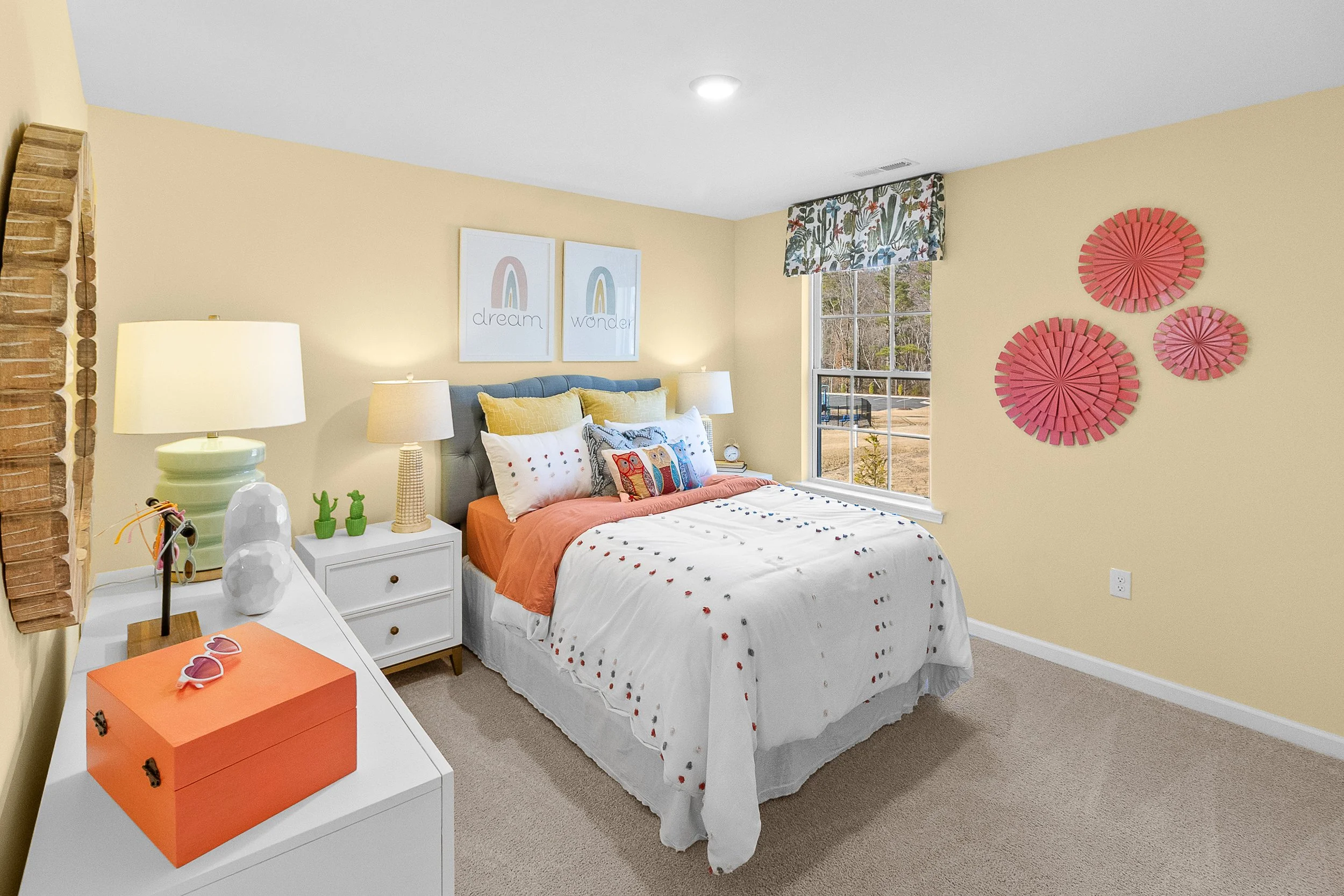 A bedroom with yellow walls, decorated with framed art, pink wall art, and a window with a patterned valance. The bed has white and yellow pillows, an orange bedspread, and a gray headboard. There are lamps on the suggests and decorative items on the