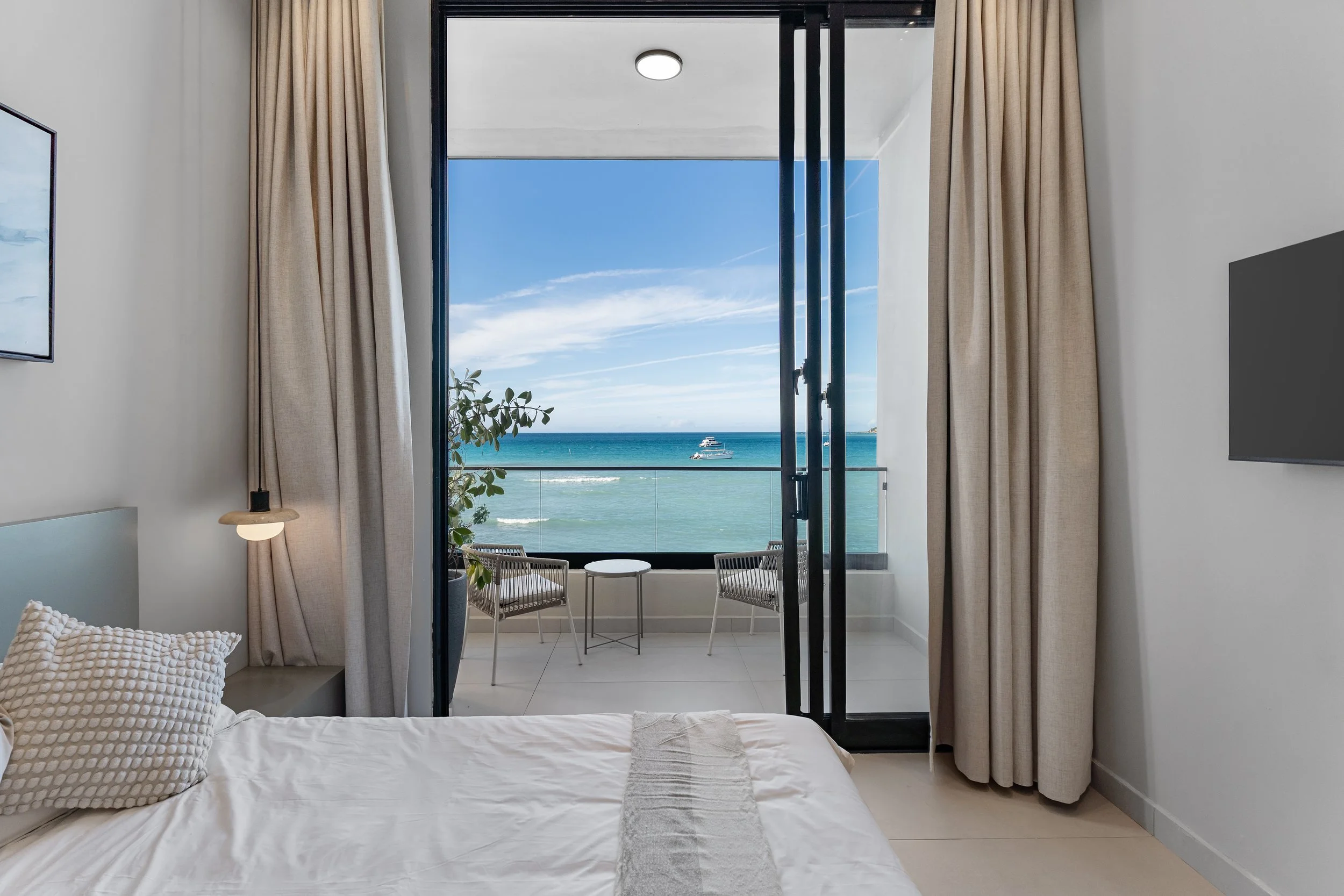 A hotel room with a bed, a nightstand, and a balcony with ocean view, two chairs, and a small table.