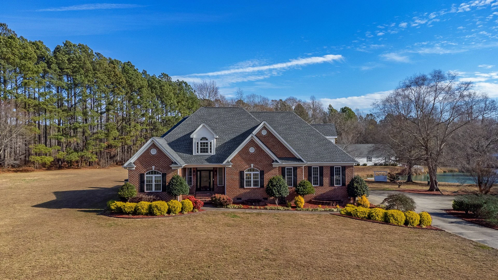 A large brick house with a front porch, surrounded by well-maintained landscaping and trees, with a forested area and a lake in the background under a mostly clear blue sky.