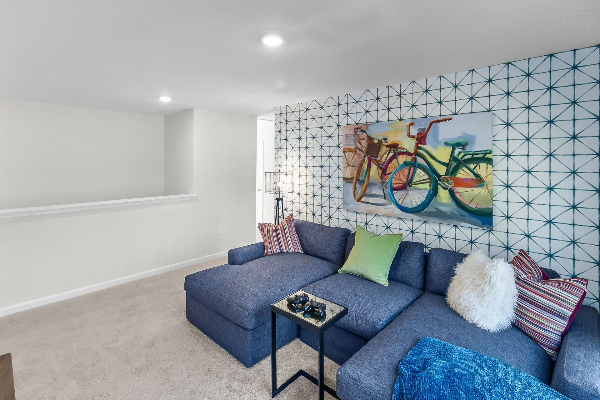Living room with white walls, a blue sectional sofa with colorful pillows, a small black side table with game controllers, a geometric wallpaper accent wall, and a vibrant painting of bicycles.