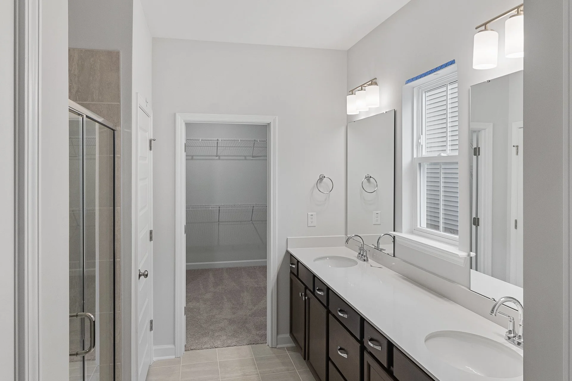 Bathroom with double vanity sinks, large mirror, window with blinds, glass shower enclosure, and an open closet with wire shelving.