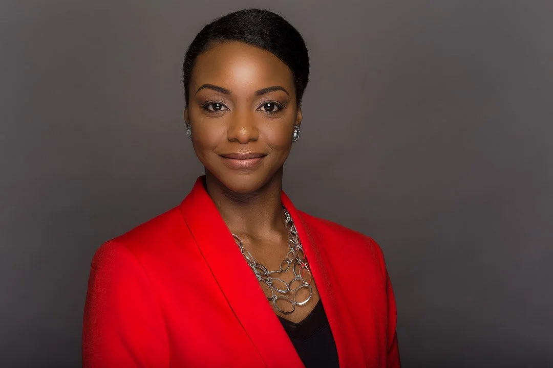 A professional woman with short black hair, wearing a red blazer, silver necklace, and earrings, smiling against a gray background.