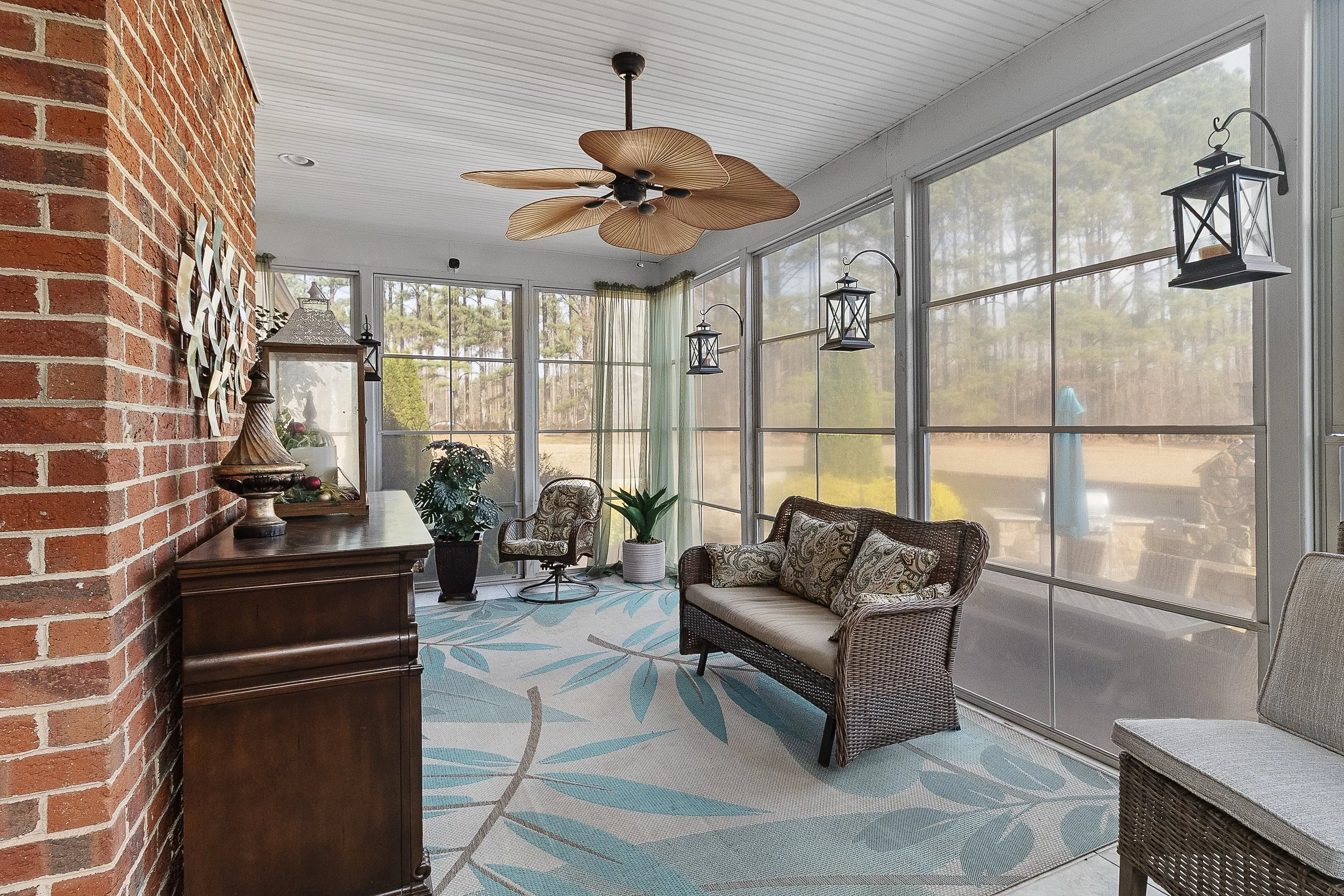 Sunroom with wicker and patterned furniture, potted plants, brick wall, ceiling fan, lanterns, and large screened windows overlooking a backyard with trees.