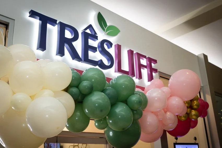 Decorated entrance with a sign that reads 'TRÈS LIFE' with green leaves above the text, and colorful balloons in cream, green, pink, gold, and red forming an arch around the sign.