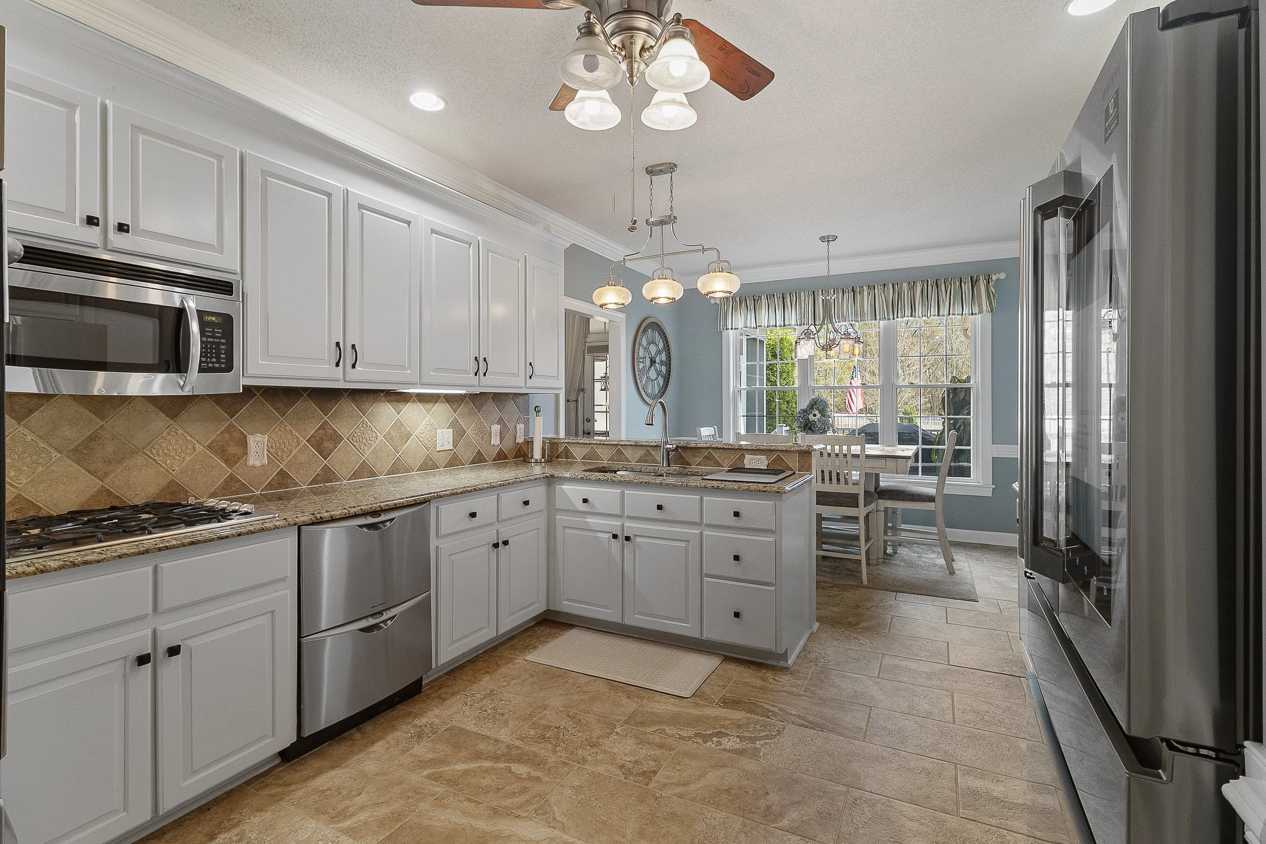 A bright, spacious kitchen with white cabinets, a granite countertop, and stainless steel appliances. There is a window with a striped valance over a dining area with a table and chairs. Overhead lighting fixtures hang from the ceiling, and a ceiling