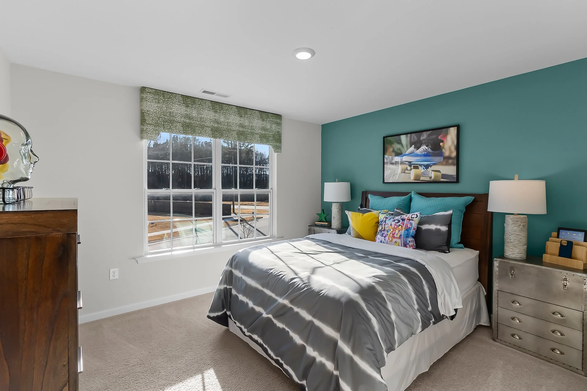 A bedroom with a bed against a teal accent wall, various colorful pillows, two nightstands with lamps, a window with a green valance, a dresser with a head bust sculpture, and a wall-mounted TV showing a skateboard.