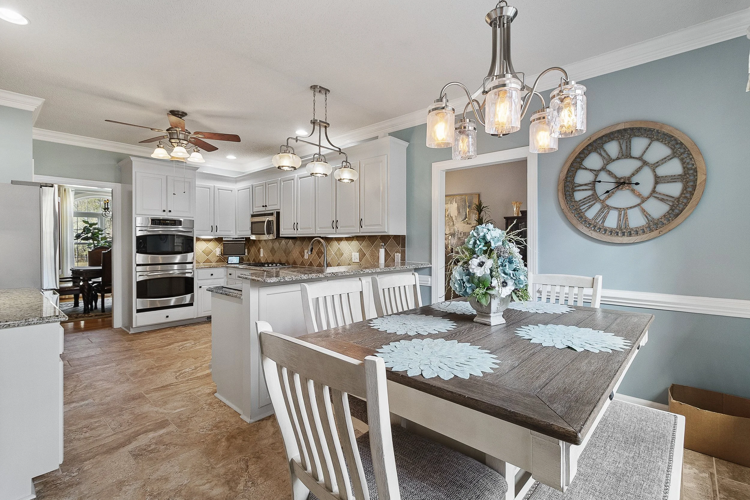 A spacious kitchen and dining area with white cabinets, granite countertops, a double oven, and a wooden dining table with four chairs. A floral centerpiece and a large wall clock are visible.