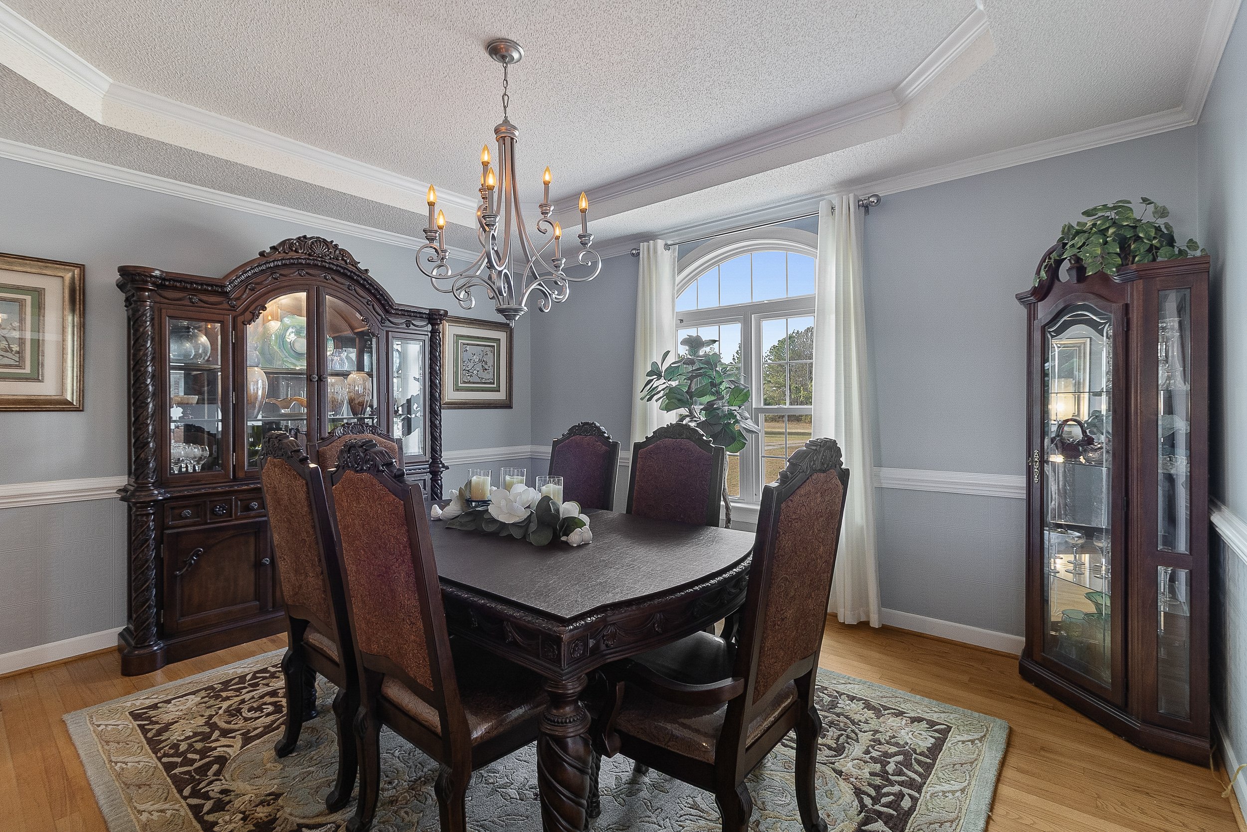 A traditional dining room with a dark wood table and six matching upholstered chairs. There's a wooden china cabinet and a glass display cabinet, both holding decorative items. A chandelier hangs above the table. A large window with white curtains le