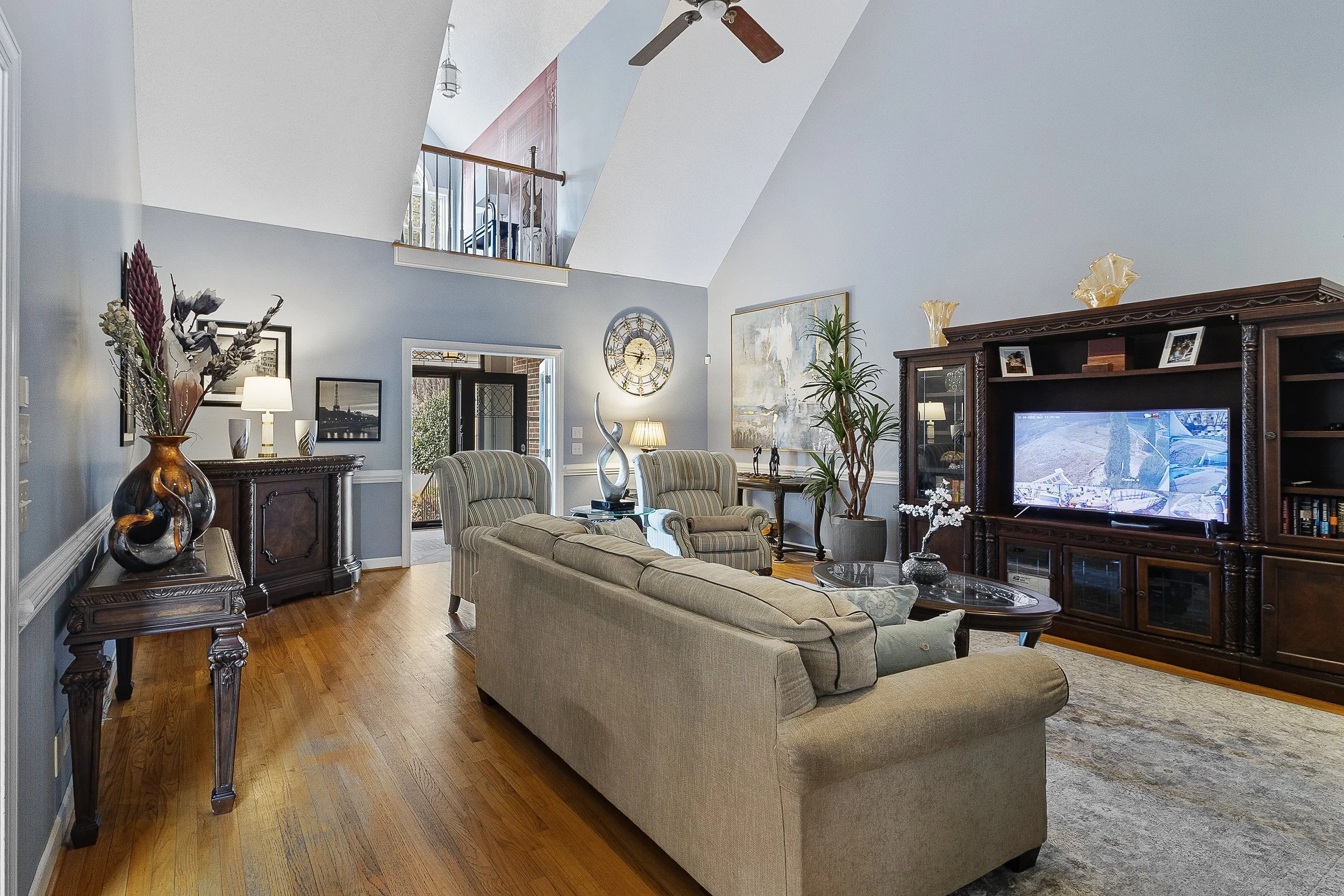 Living room with beige sofa, striped armchairs, dark wood entertainment center with TV, decorative vases, large painting, floor lamp, ceiling fan, hardwood floors, and front door in background.