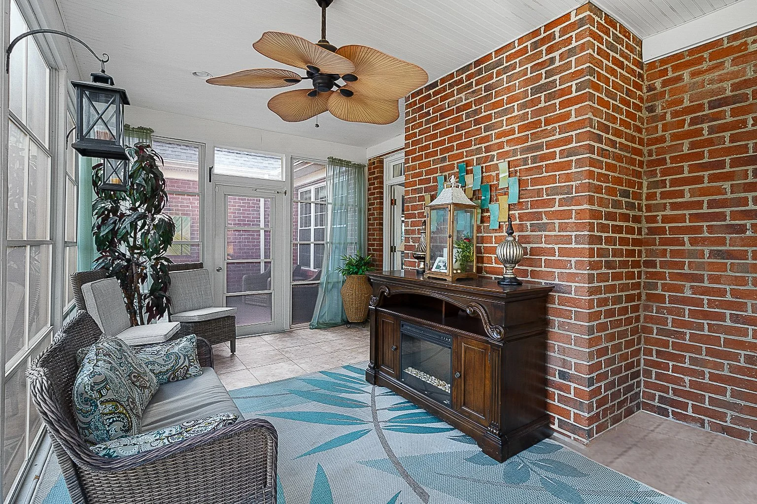 Screened-in porch with wicker furniture, potted plant, brick wall, wooden console table, and decorative items, including a ceiling fan with leaf-shaped blades.