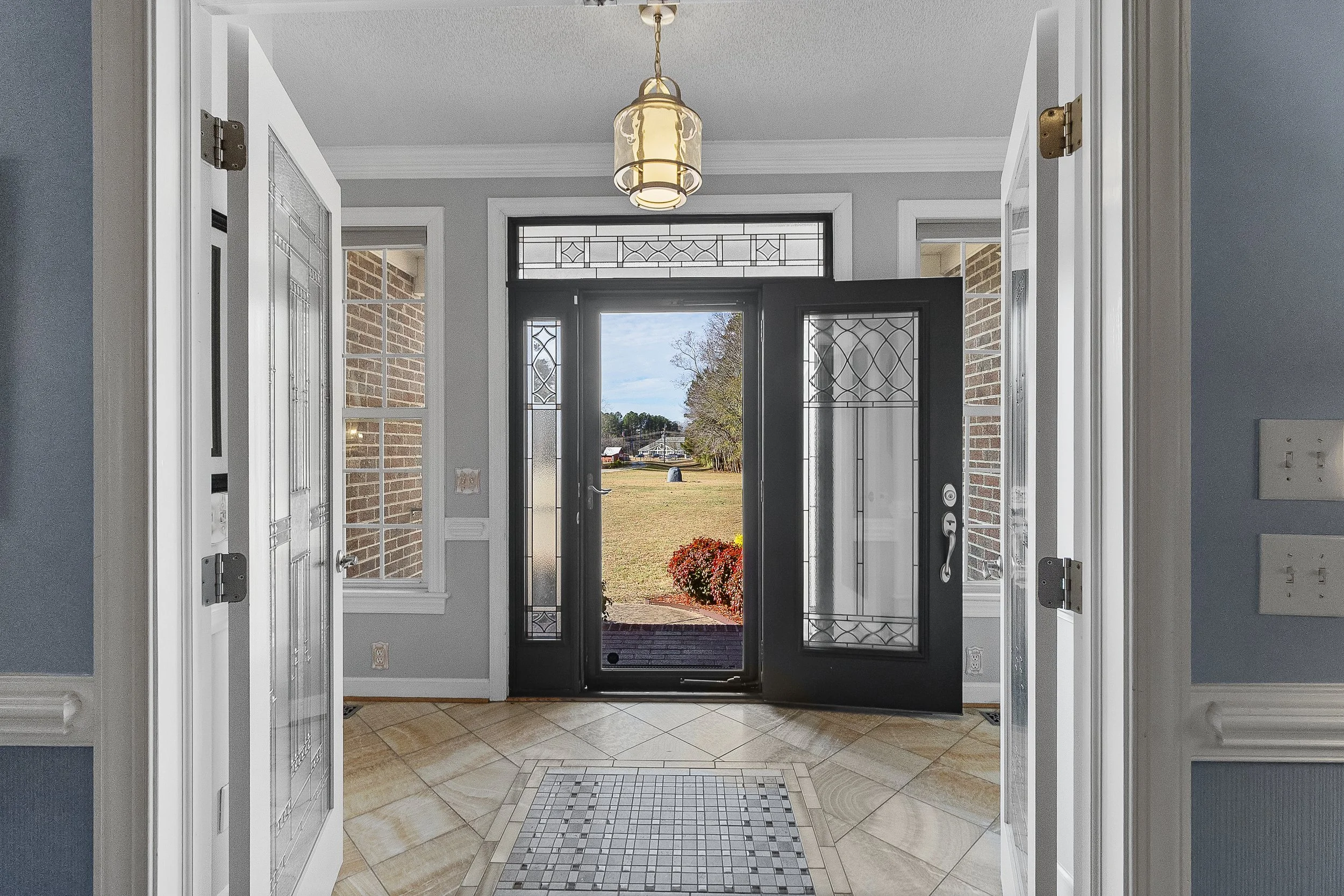 Foyer of a house with an open black front door, decorative glass panels, and a view of the yard outside. The interior has light gray walls, white trim, a tiled floor, and a hanging light fixture.