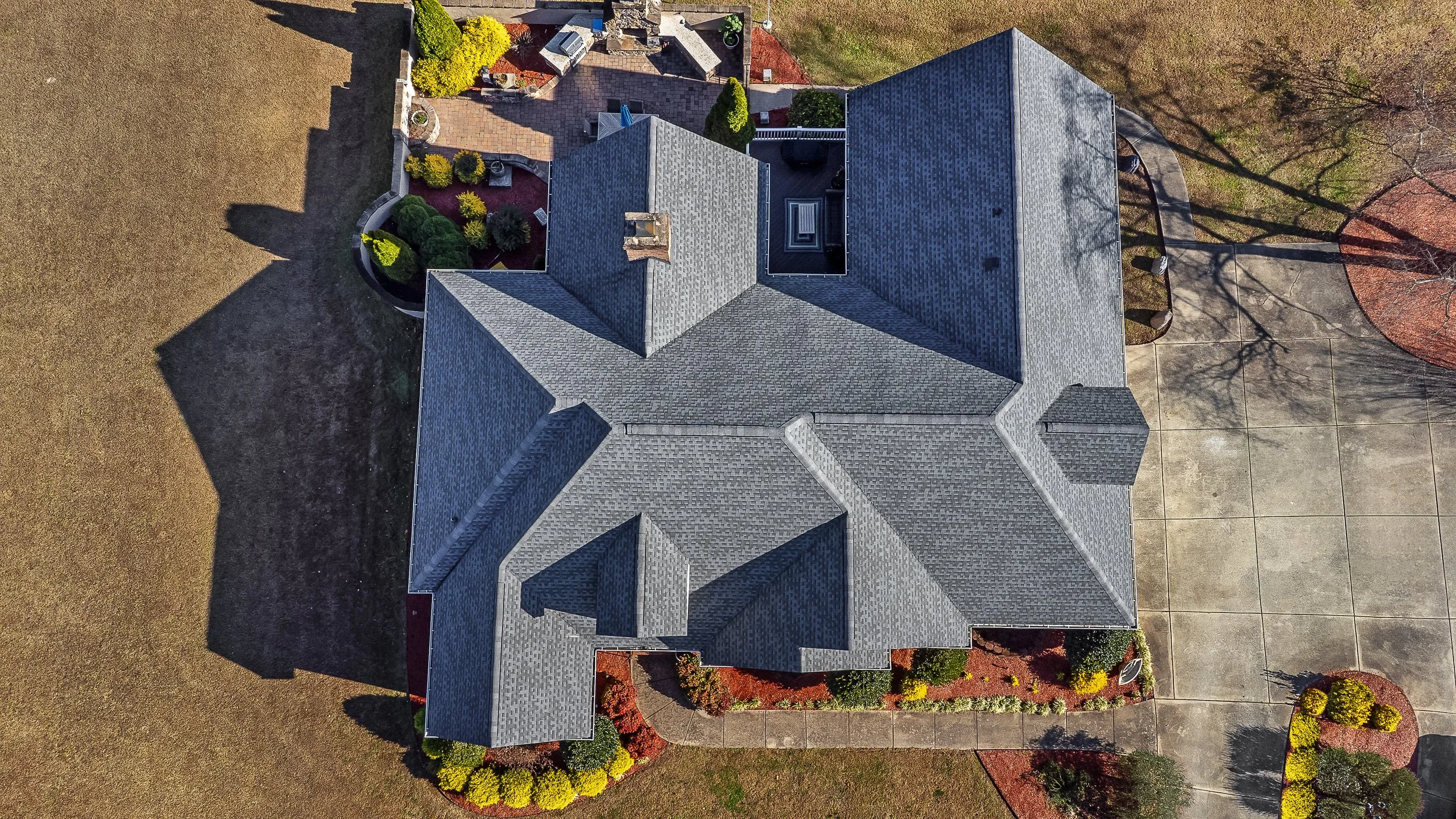 An aerial view of a large residential house with a gray shingle roof, surrounded by a landscaped yard with flower beds, trees, and a driveway.