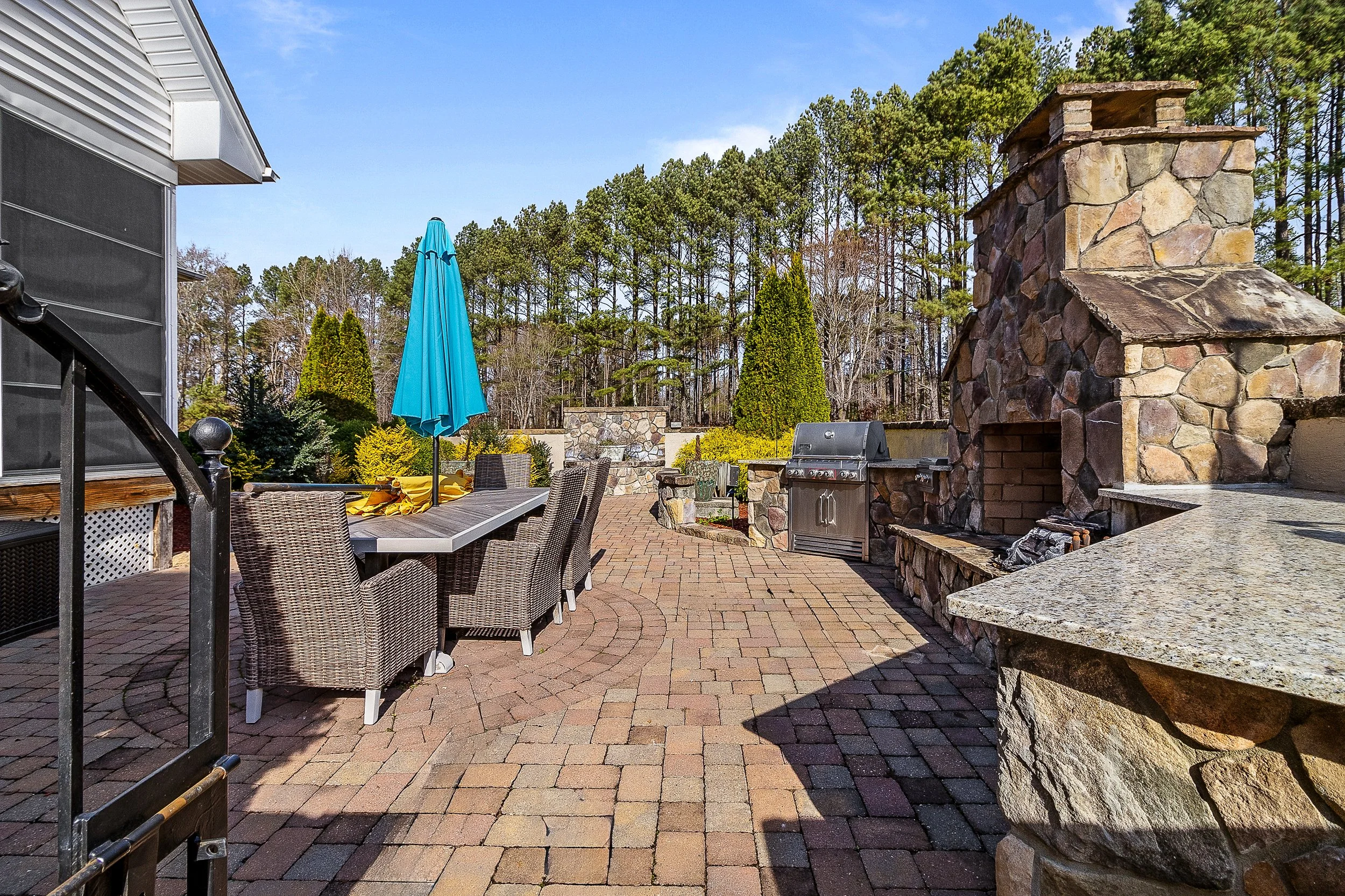 Outdoor patio area with brick pavers, stone fireplace, outdoor grill, patio furniture, umbrella, and a wooded backyard.