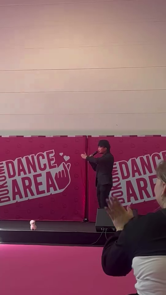 Junger Mann in schwarzem Anzug macht Selfie in einer modernen, hellen Eingangshalle mit Glas-Türen und einer Person im Hintergrund, die 스포츠트크le trägt.