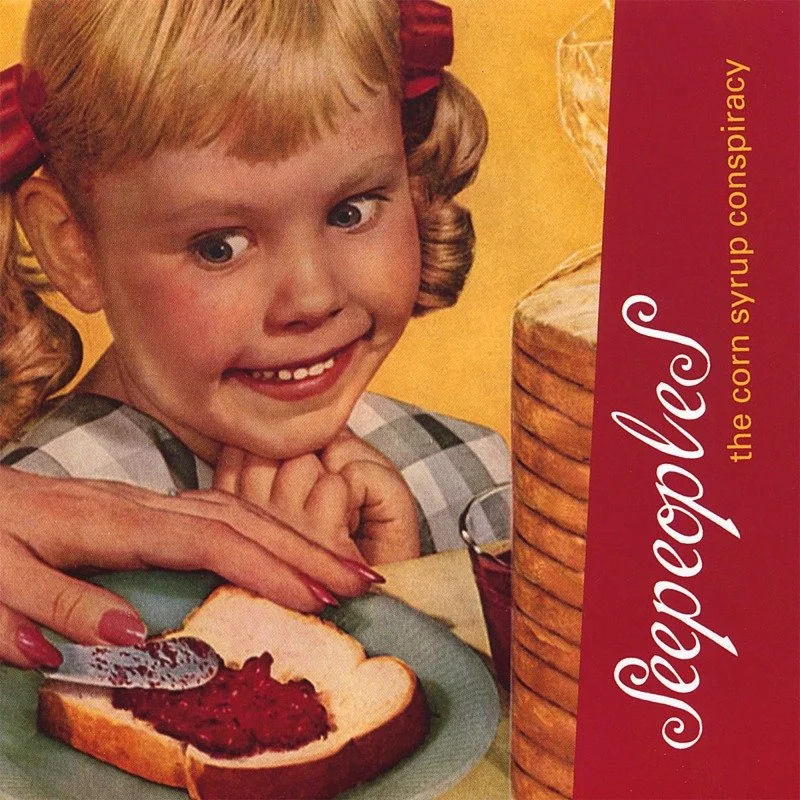 The image shows a young girl with blonde hair in pigtails, smiling and resting her chin on her hands. In front of her, an adult's hand is spreading corn syrup on a slice of bread with raspberry jam. To the right, there is a stack of round cookies or crackers and a red box of