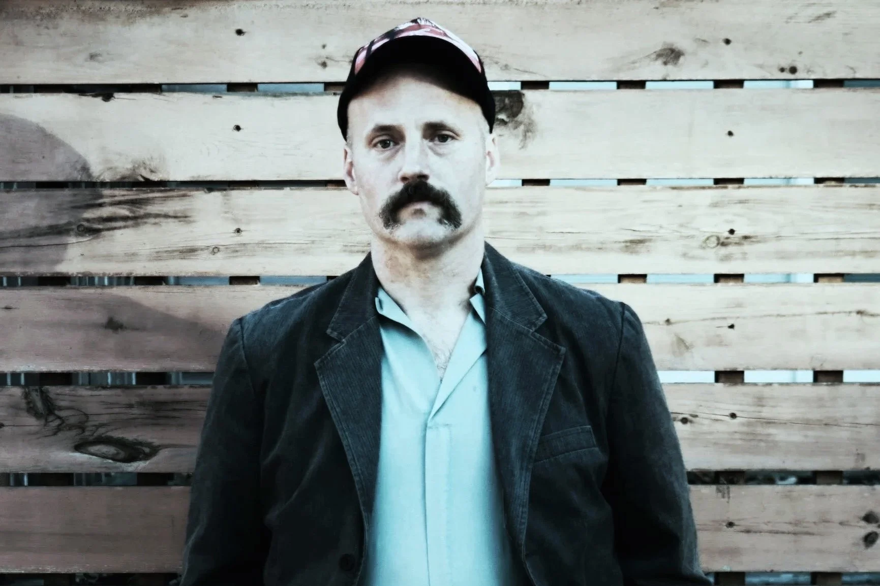 A man with a mustache and beard wearing a black jacket, light shirt, and a cap, standing in front of a wooden fence with horizontal planks.