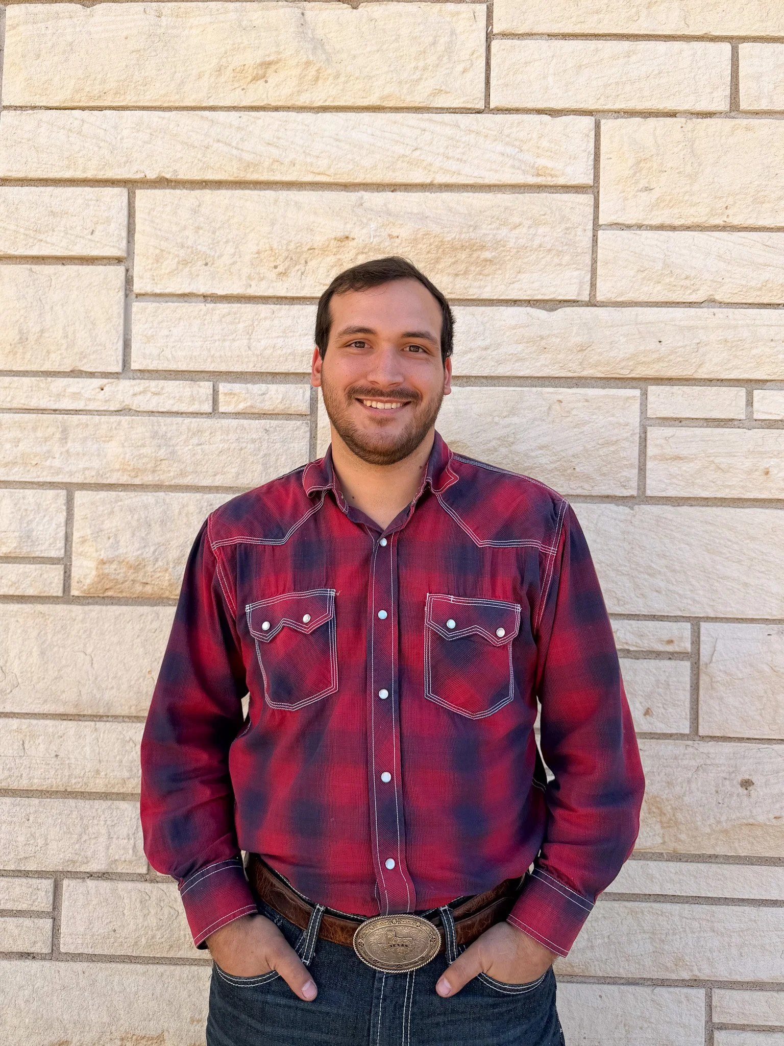 A man with brown hair and a beard smiling, wearing a red and black plaid shirt with white stitching, standing in front of a beige brick wall.