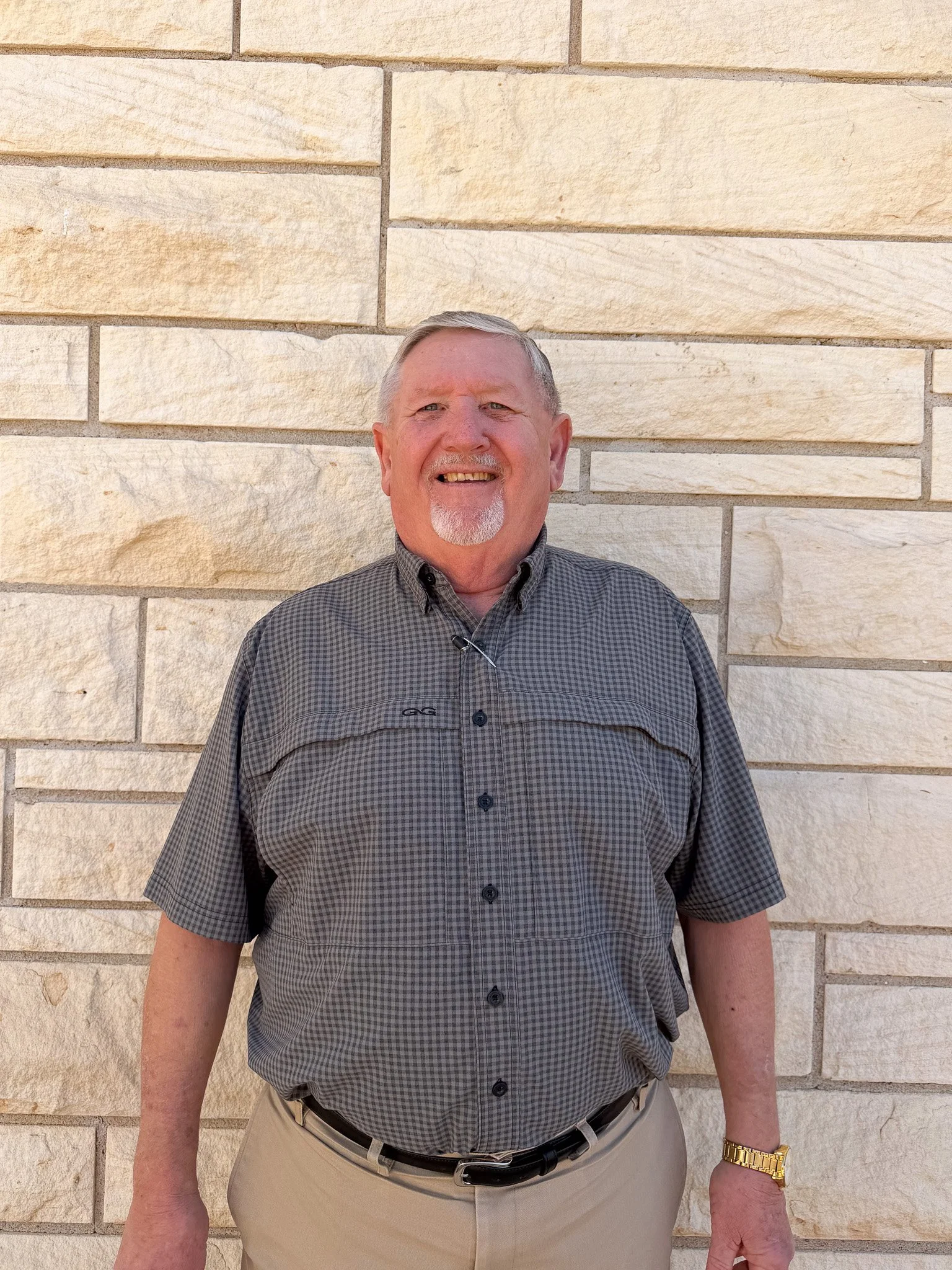 A smiling man standing in front of a beige brick wall, wearing a gray checkered short-sleeve shirt, beige pants, and a gold watch.