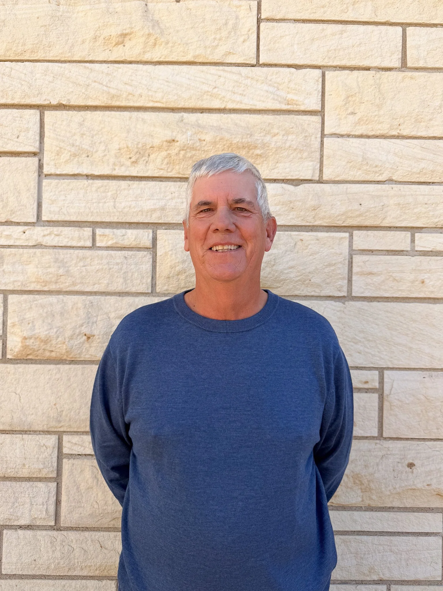 A smiling man with gray hair standing in front of a beige stone wall, wearing a navy blue long-sleeved shirt.