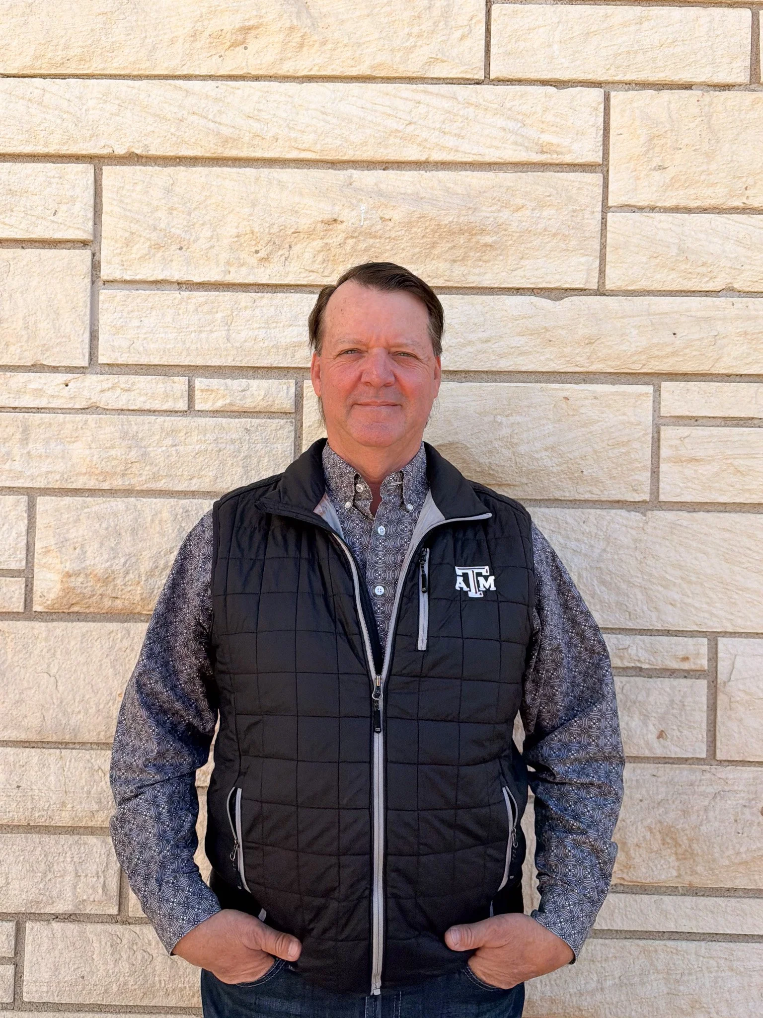 A man stands in front of a beige brick wall, wearing a black Texas A&M vest over a patterned shirt, with hands in his pockets and a neutral expression.