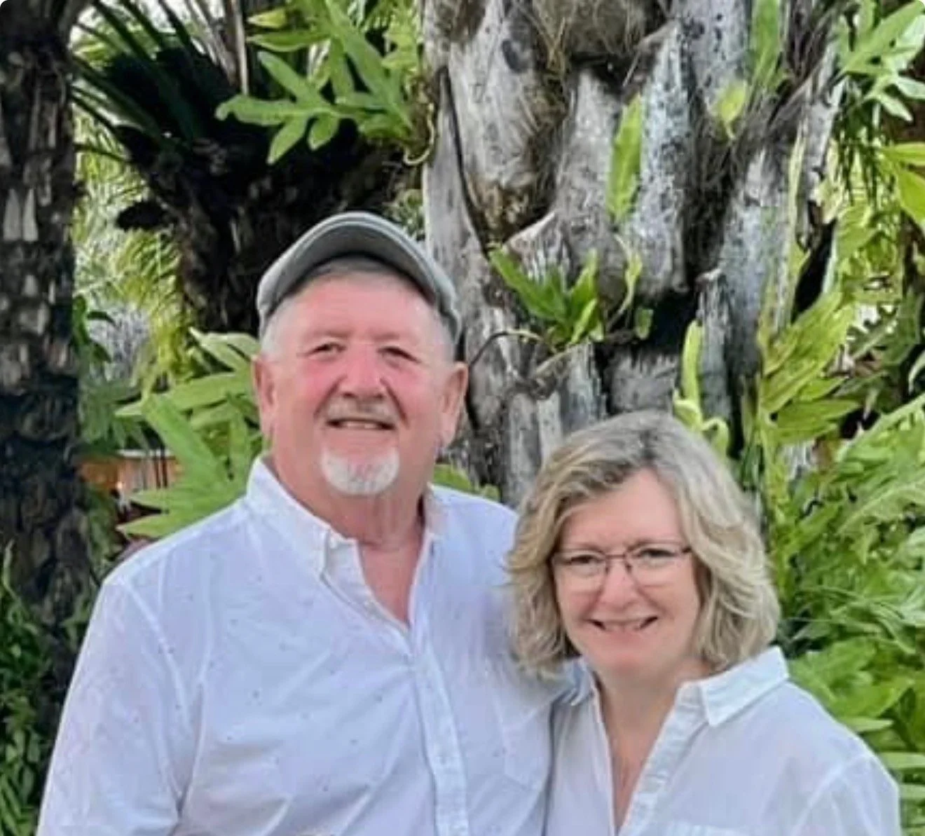 A smiling man and woman in white shirts standing outdoors in front of a tree with green leaves.