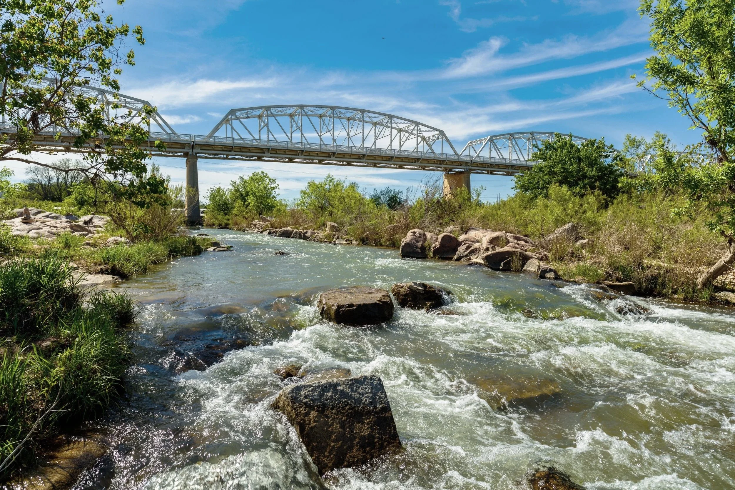 A river with rocks flowing under a bridge on a sunny day with blue sky and green trees.