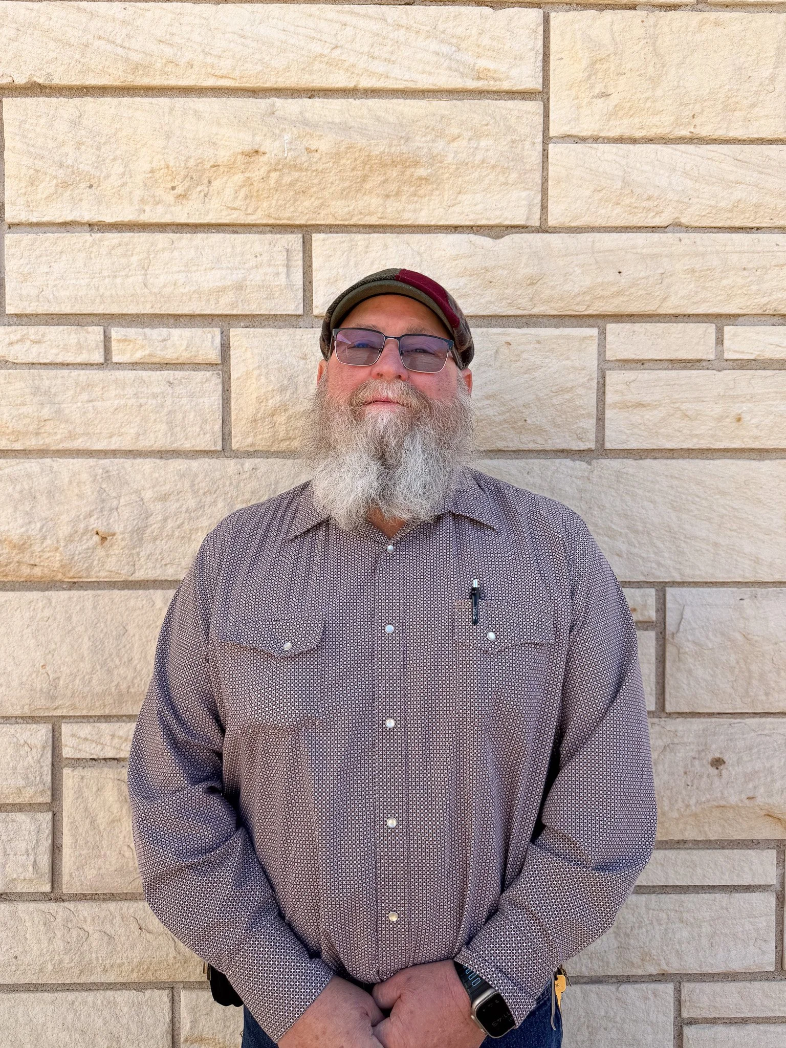 A man with a beard and glasses standing against a beige brick wall.
