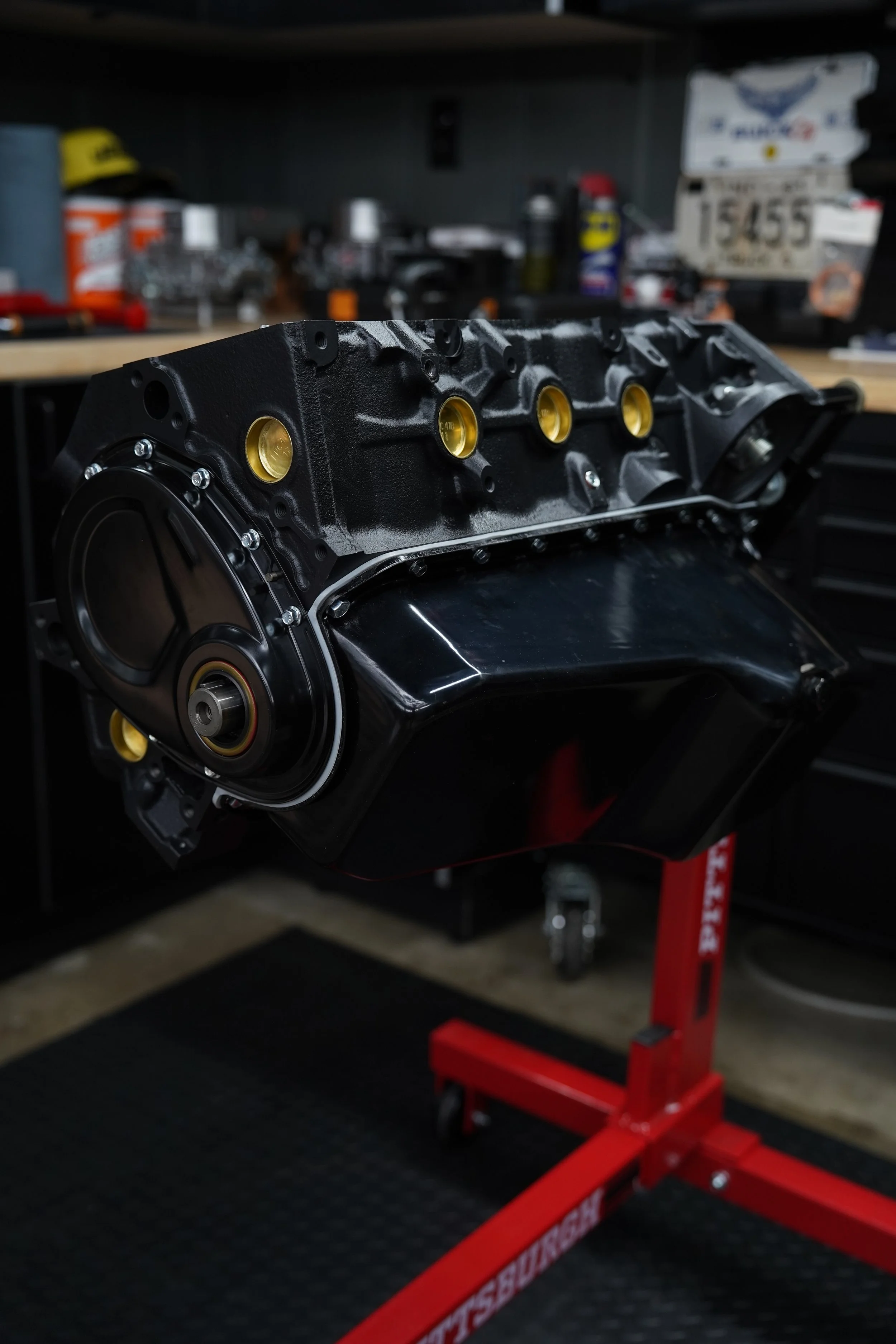 Automotive engine block on a red stand in a workshop with tools and supplies on shelves in the background.