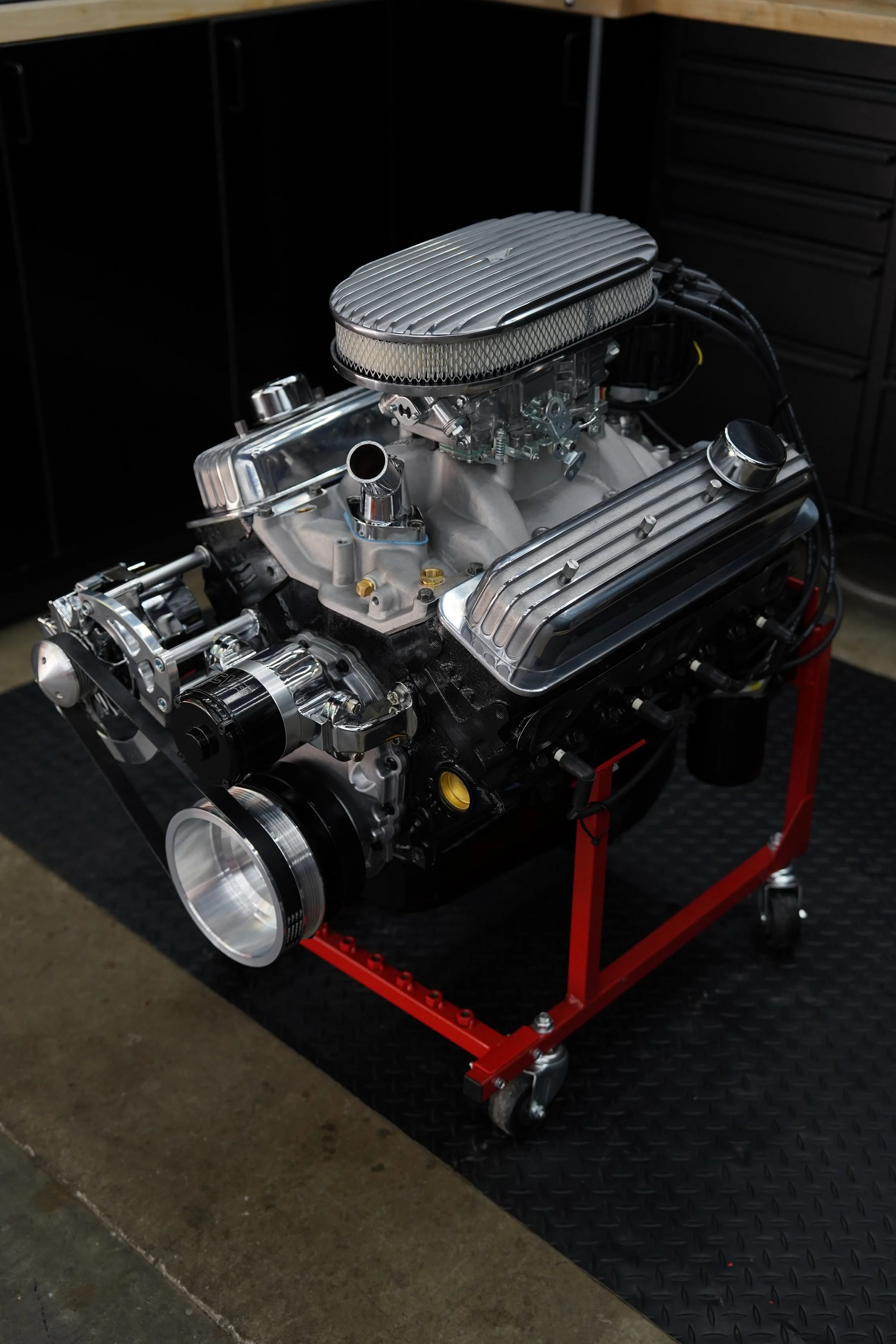 A high-performance V8 engine on a red engine stand with black caster wheels in a workshop. The engine has shiny chrome components, a large blake air filter, and aluminum valve covers.
