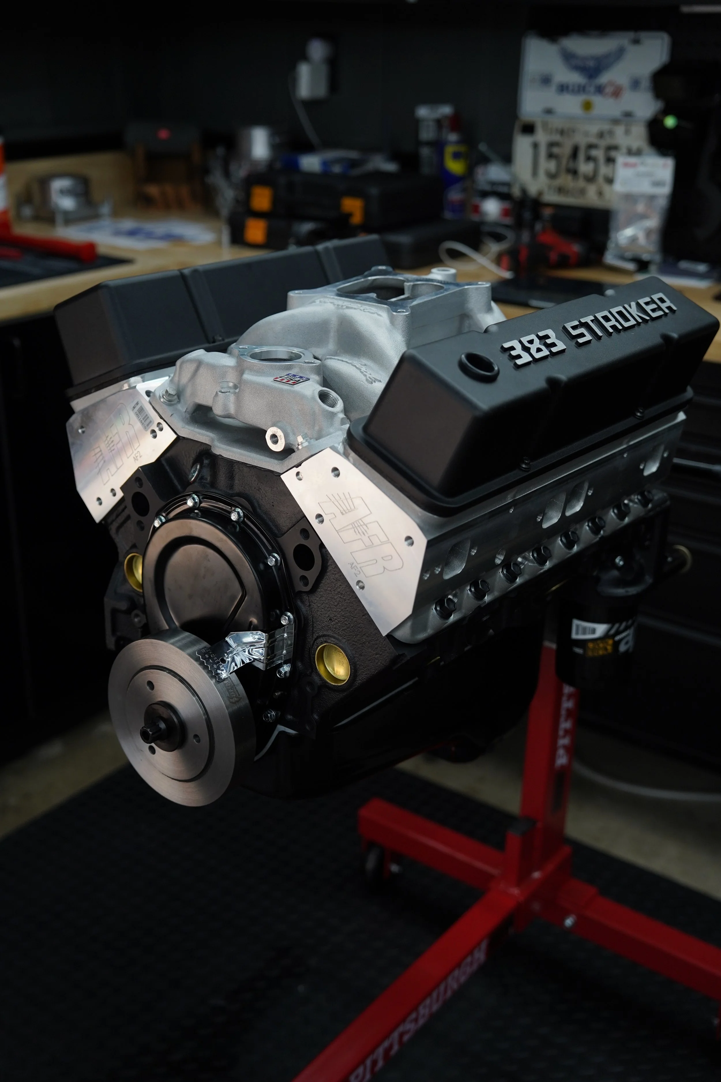 New automotive engine with black valve covers labeled '383 STROKER' on a workbench in a garage or workshop.