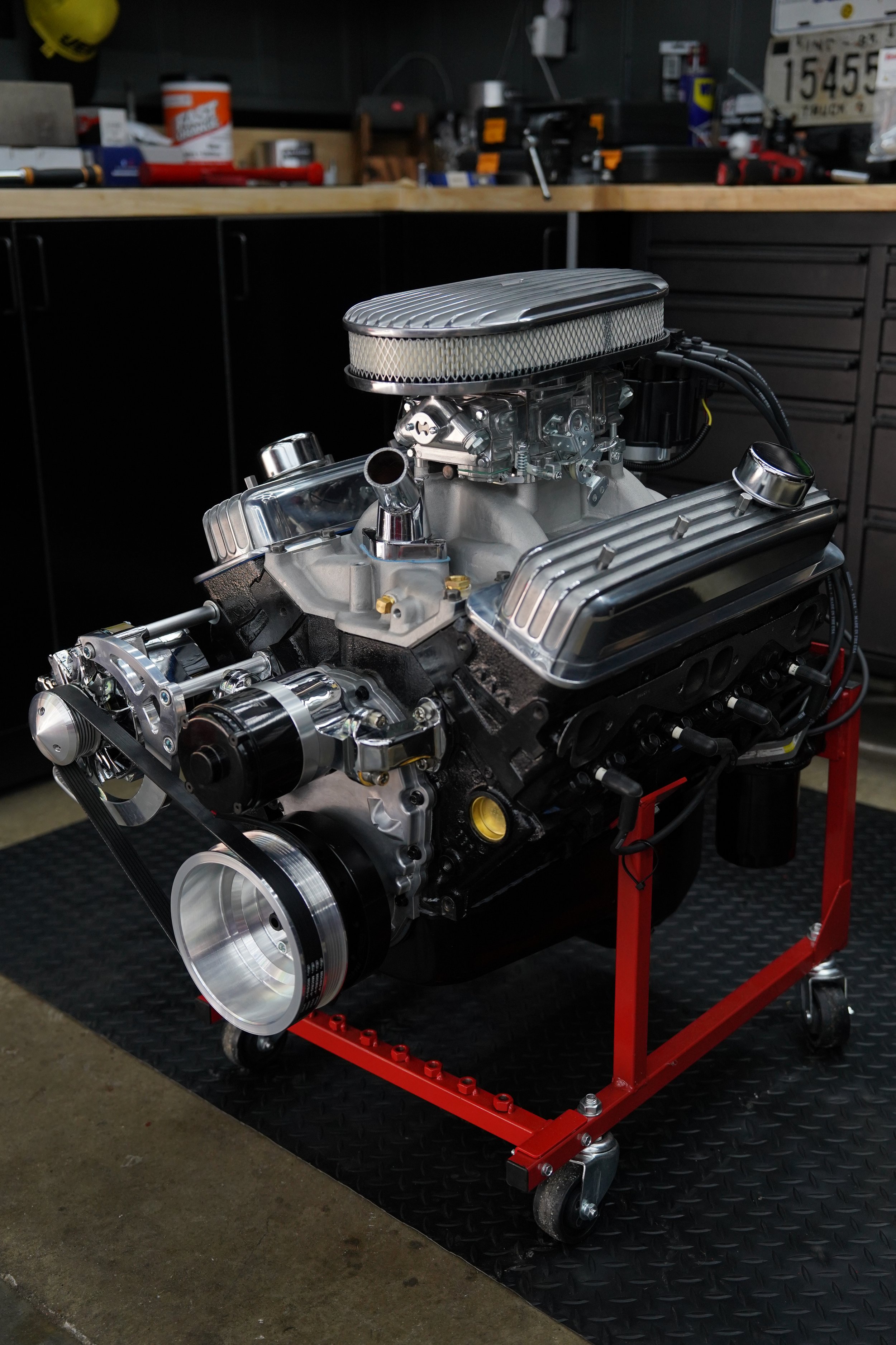 A chrome and black V8 engine on a red engine stand in a workshop with tools and parts in the background.