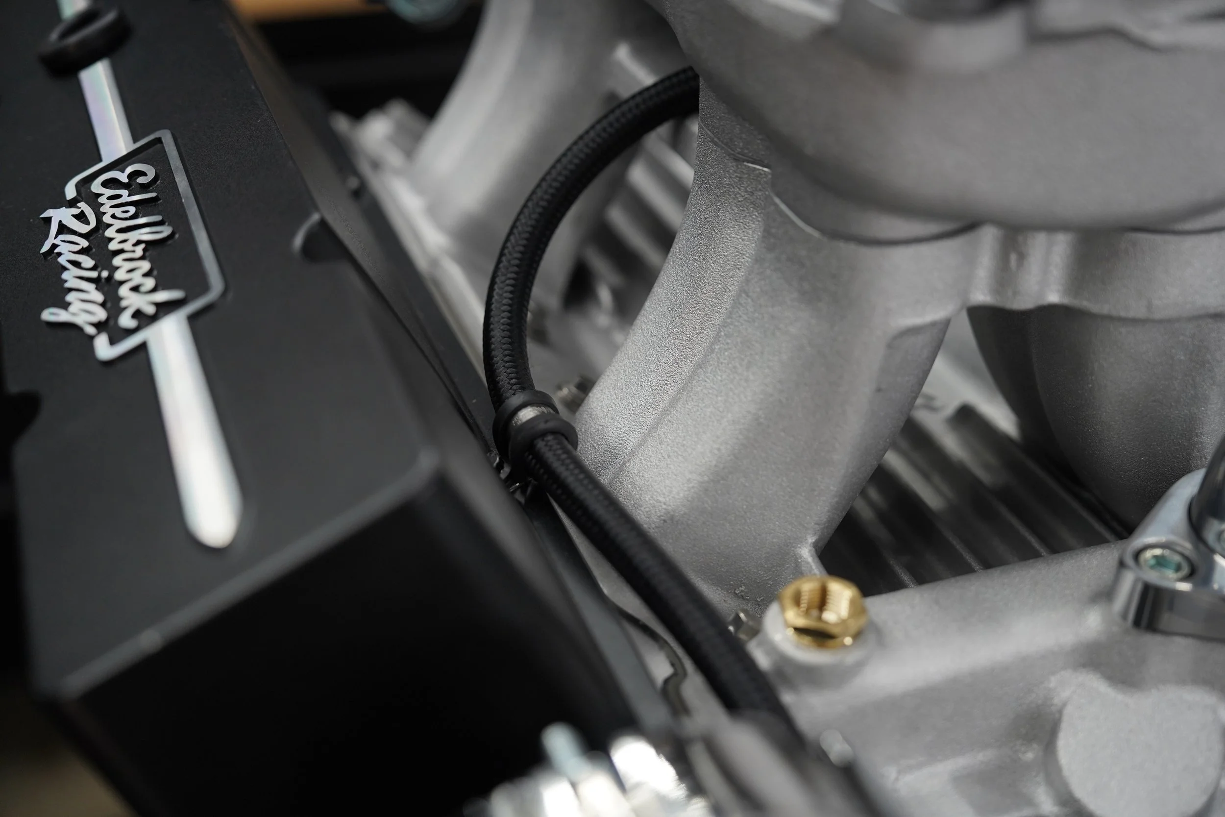 Close-up of a metallic engine component with a black cover labeled 'Wilwood'. A black cable runs across the component, which has a textured surface and several bolts.