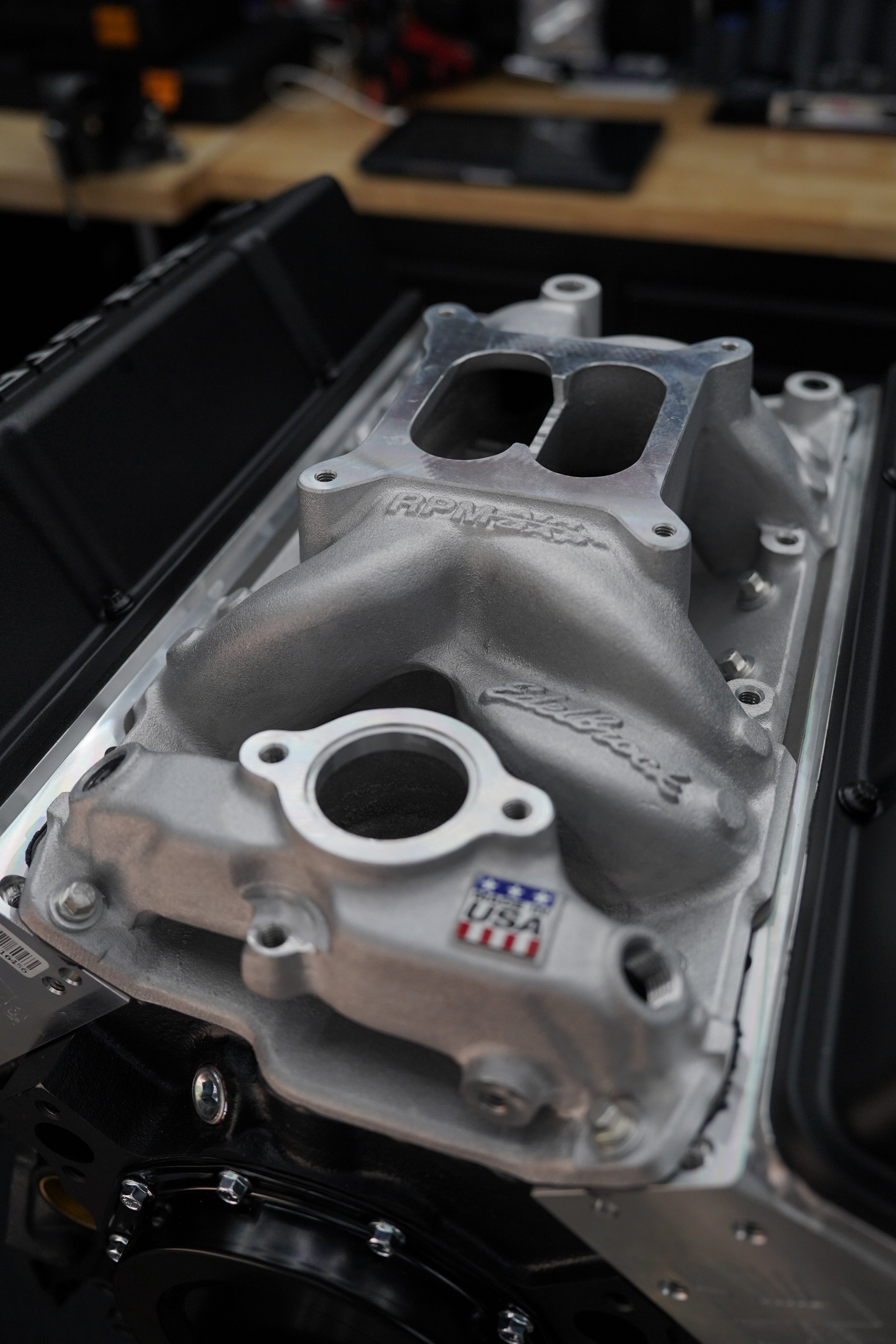 Close-up of a car engine component, featuring a metallic intake manifold with branding, on a workbench with tools in the background.