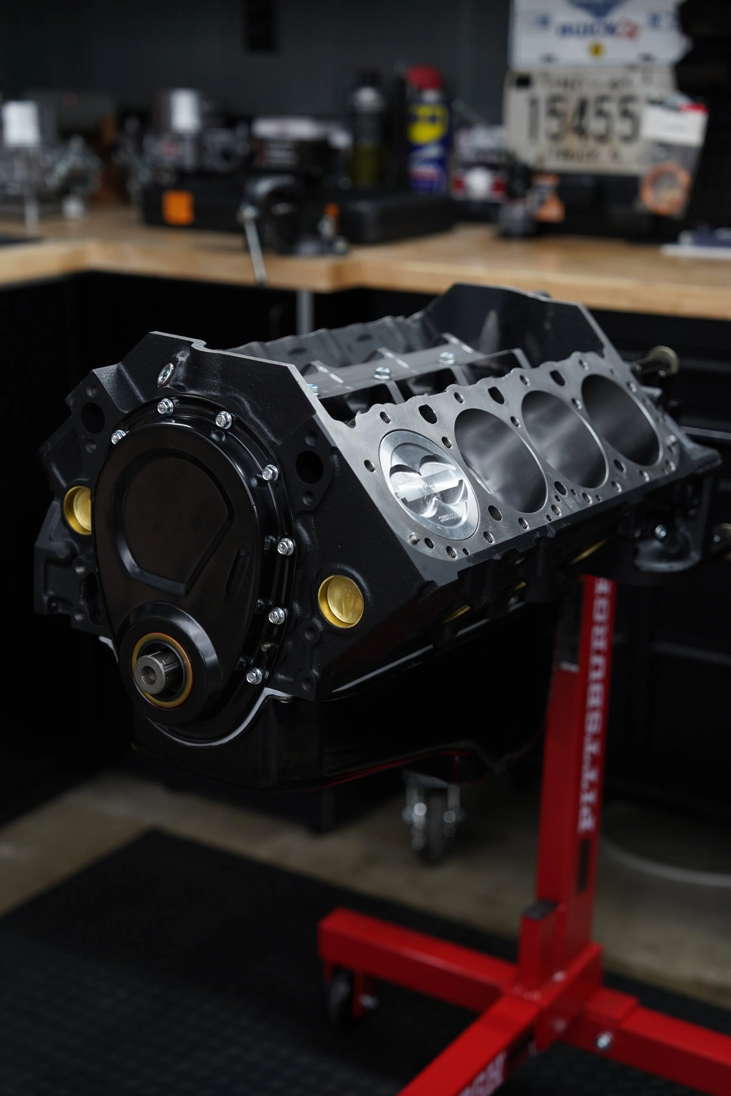 A car engine block mounted on a stand in a workshop with tools and parts in the background.