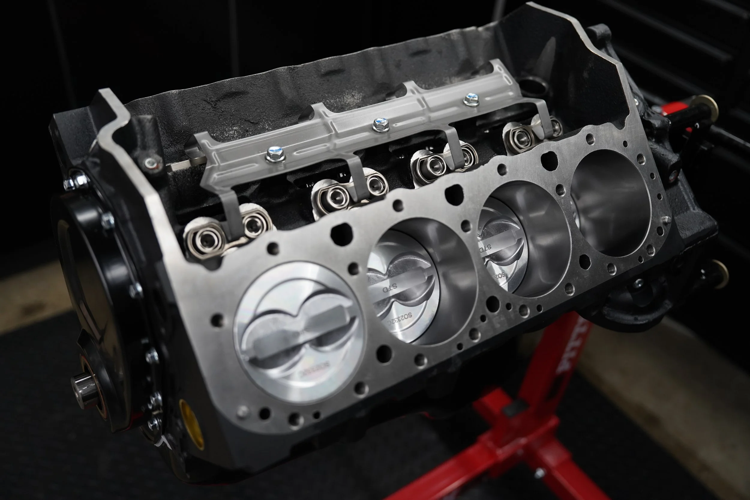 Close-up of a car engine block with pistons, valves, and camshaft components visible, mounted on a red engine stand.