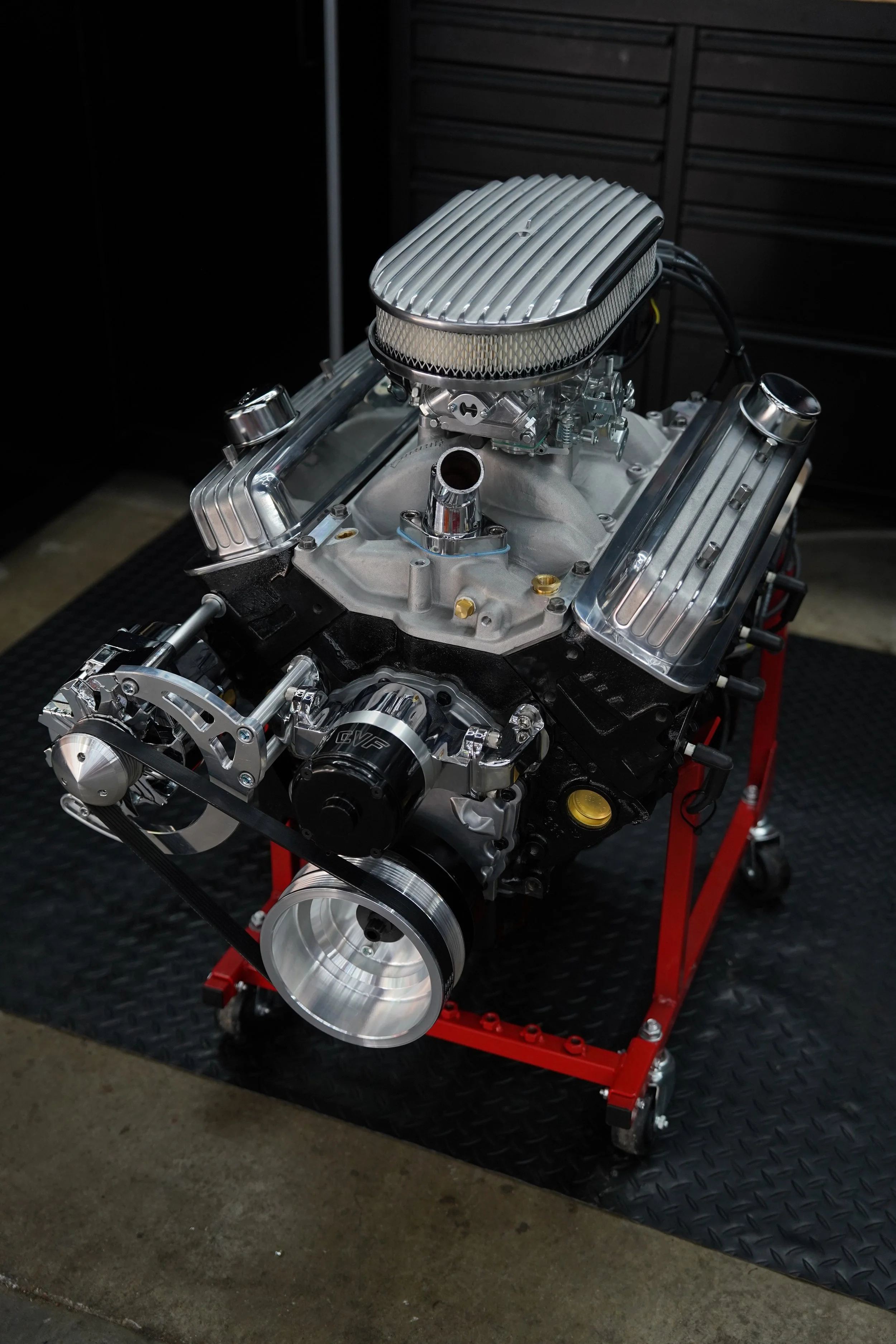 A car engine with a polished intake manifold and valve covers on a black workbench with a red frame, in a workshop setting.